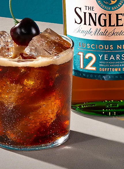 A whisky cocktail with a cherry on top next to a bottle of The Singleton 12 Year Old Single Malt Scotch Whisky.