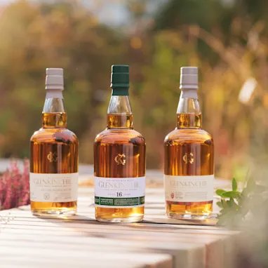 Een vrouw met een turkooizen trui glimlacht terwijl ze zit en een glas met ijs en whisky vasthoudt.