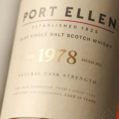 De witte gebouwen van de Port Ellen distilleerderij kijken uit over een waterpartij omgeven door een rotsachtige kust. Op een van de witte muren staan in grote zwarte letters de woorden 'Port Ellen'.