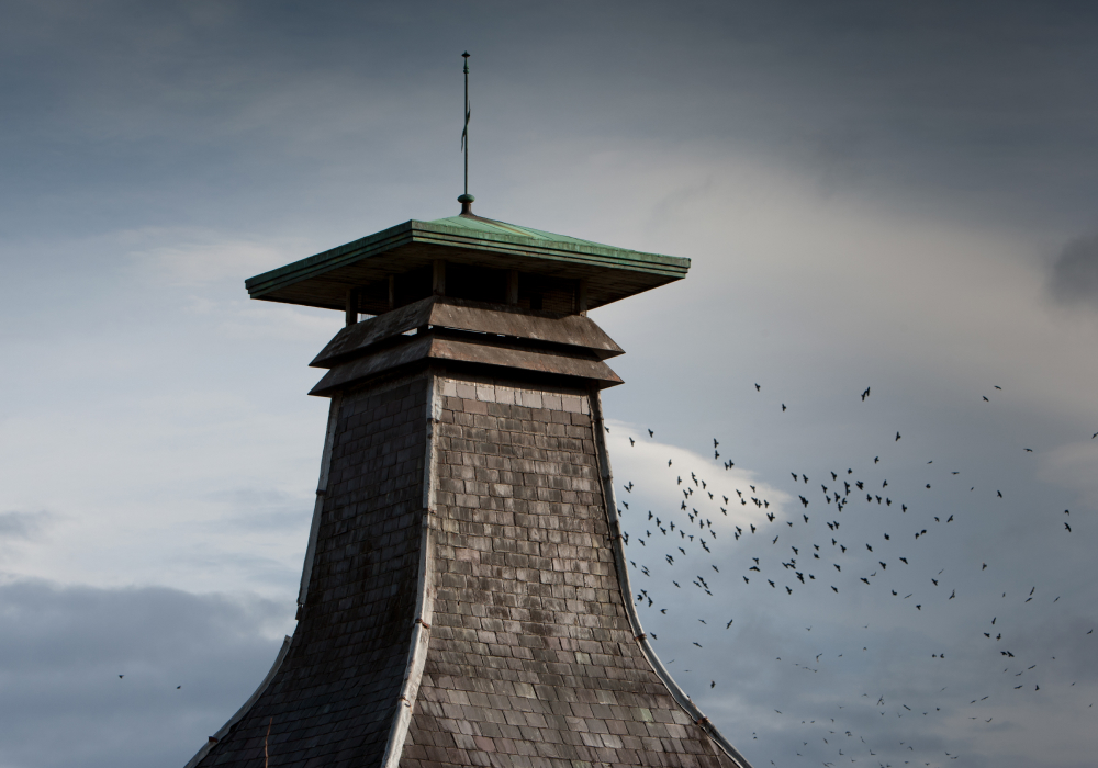 Een schoorsteen in pagodestijl staat tegen een blauwe lucht met witte wolken, met een zwerm vogels erachter.
