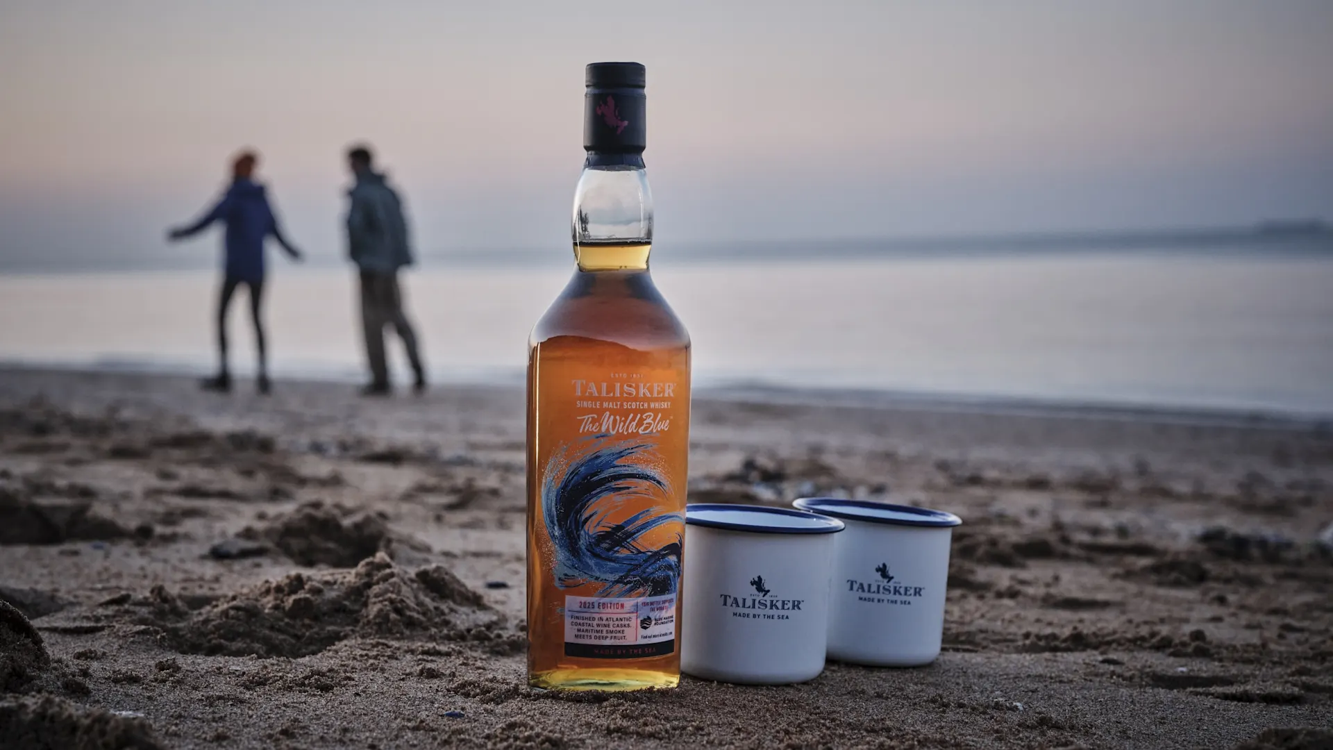 Een fles Talisker "The Wild Blue" single malt Scotch whisky staat op een zandstrand tijdens de schemering, met daarnaast twee emaille mokken met Talisker-logo. Op de achtergrond wandelen een man en een vrouw onscherp langs de kustlijn.