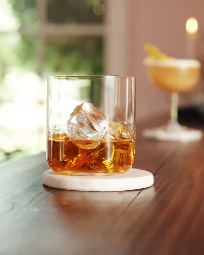 A rocks glass containing whisky and ice sits on a bar, with a cocktail shaker and jigger behind it.