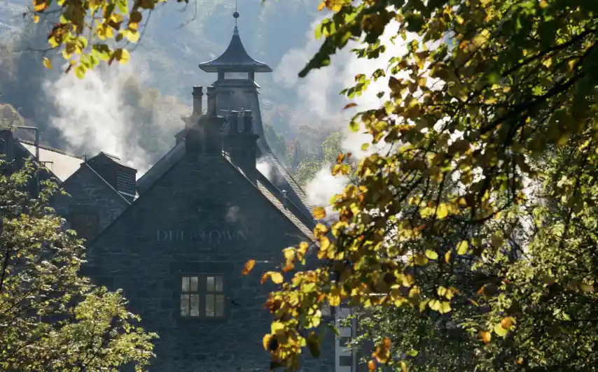 De stenen muren van de Singleton distilleerderij staan tussen twee groene bomen. De pagodeschoorsteen van een gebouw staat voor een bergketen bedekt met bomen.