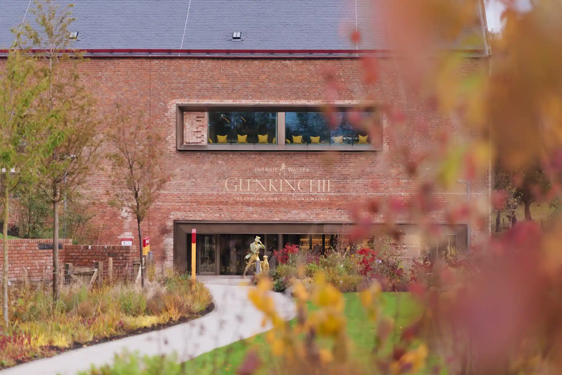 A red brick wall of Glenkinchie distillery sits at the end of a path surrounded by colourful plants. A golden Johnnie Walker ‘Striding Man’ statue is at the end of the path, and the sign on the wall says ‘Glenkinchie’.