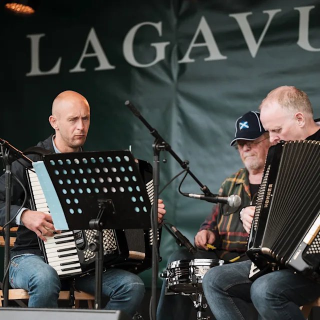 Zwei Männer mit Akkordeons und ein Mann mit einem Schlagzeug sitzen vor Mikrofonen, hinter denen die Worte Lagavulin stehen.