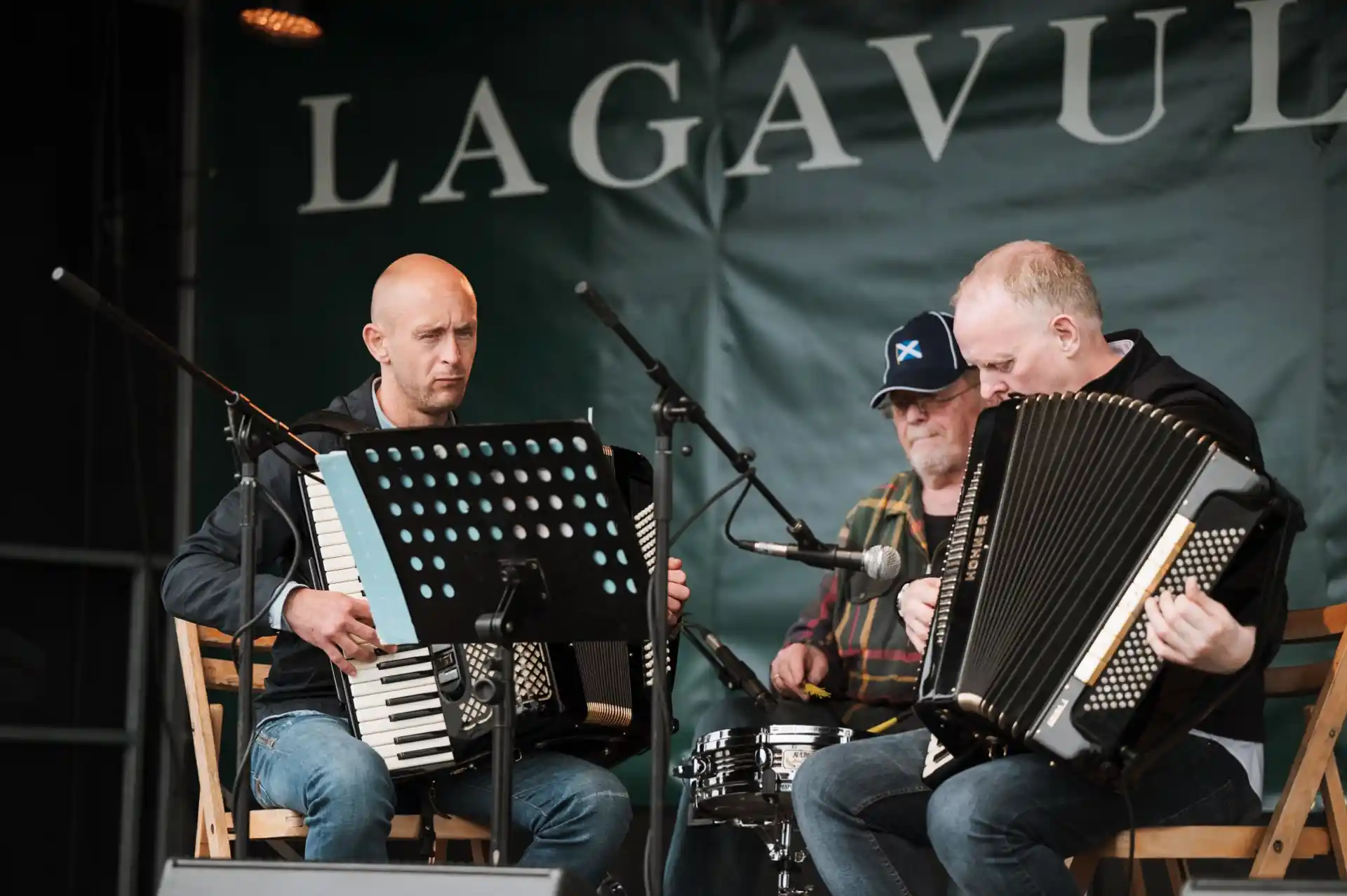 Two men with accordions and one man with a drum set sit on front of microphones with the words Lagavulin behind them.