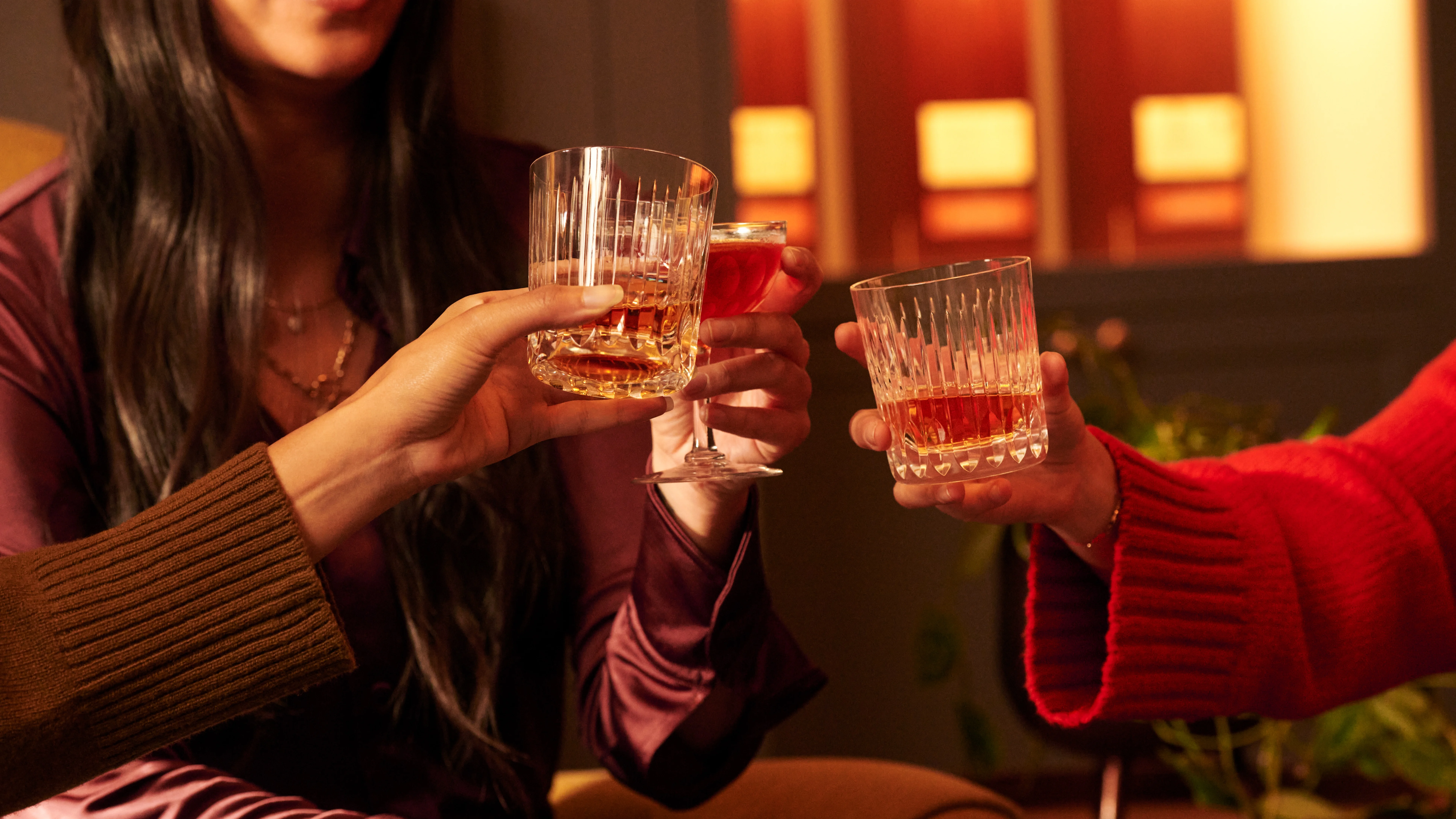 Three women hold glasses together in a 'cheers' motion. They contain assorted whisky cocktails.