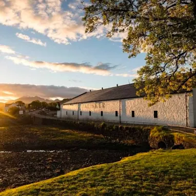 The white building of Talisker distillery sits on a green piece of land, above a pebbly shore. The shore flows into a body of water and beyond the horizon is a sunset.