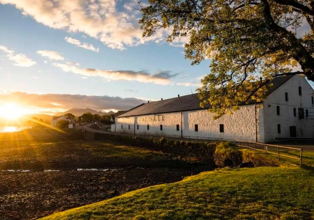 The white building of Talisker distillery sits on a green piece of land, above a pebbly shore. The shore flows into a body of water and beyond the horizon is a sunset.