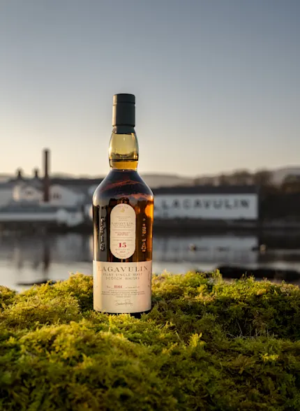 Bottle of Lagavulin Fèis Ìle 2025, sat on mossy ground in front of the distillery at sunset