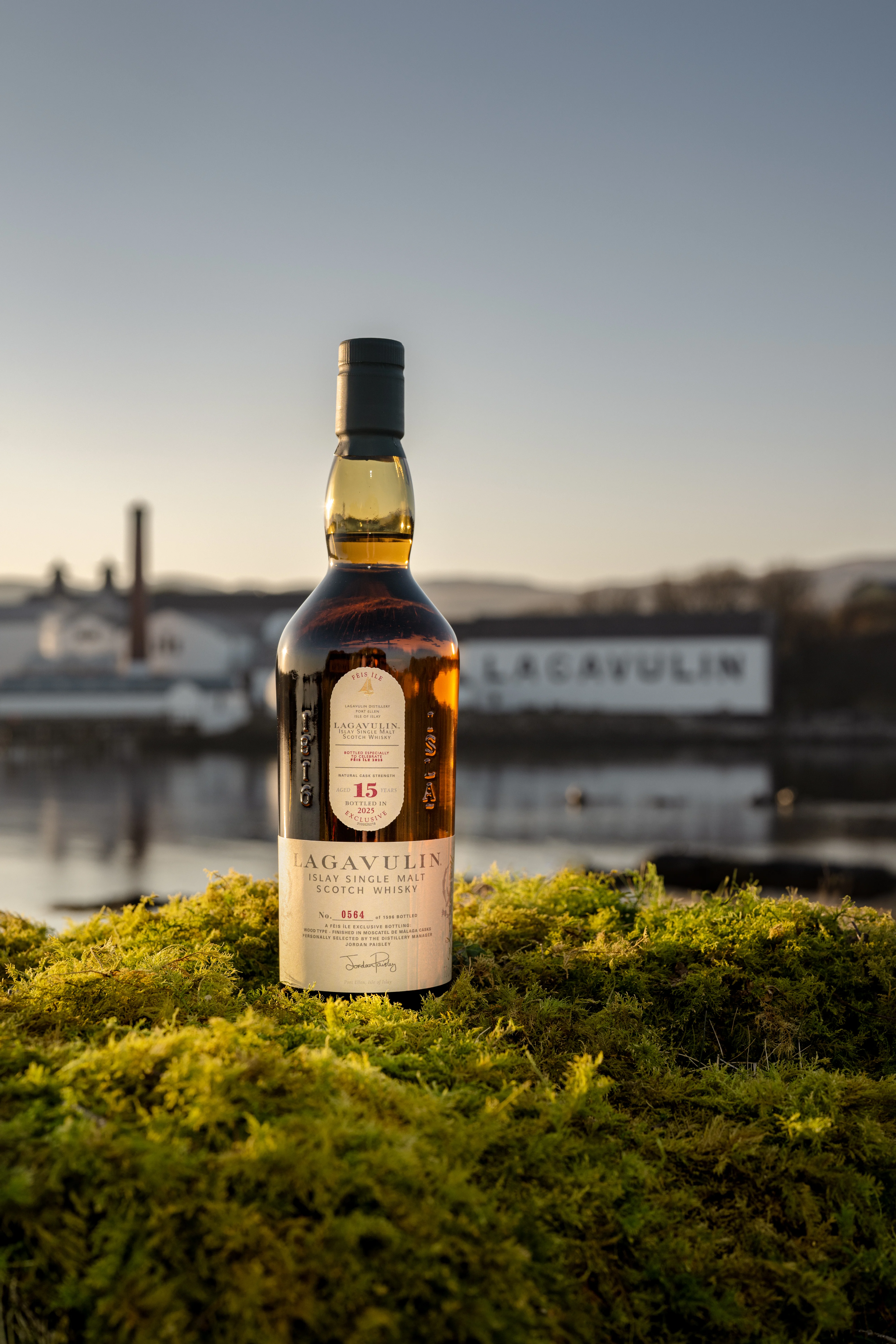 Bottle of Lagavulin Fèis Ìle 2025, sat on mossy ground in front of the distillery at sunset