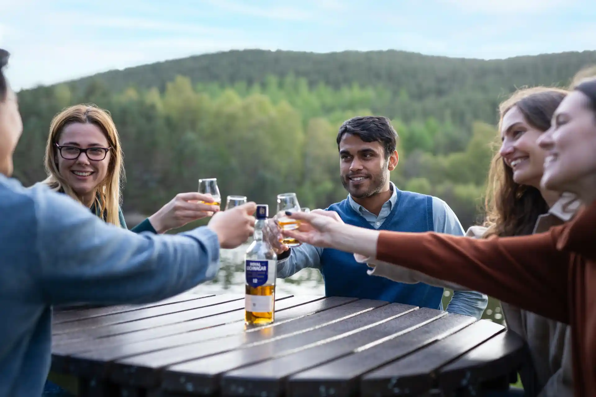 A group of people sit outside at a round table on which there is a bottle of Royal Lochnagar whisky. They each hold a dram glass of whisky and bring the glasses together to ‘cheers’. Behind them is a mountain range.