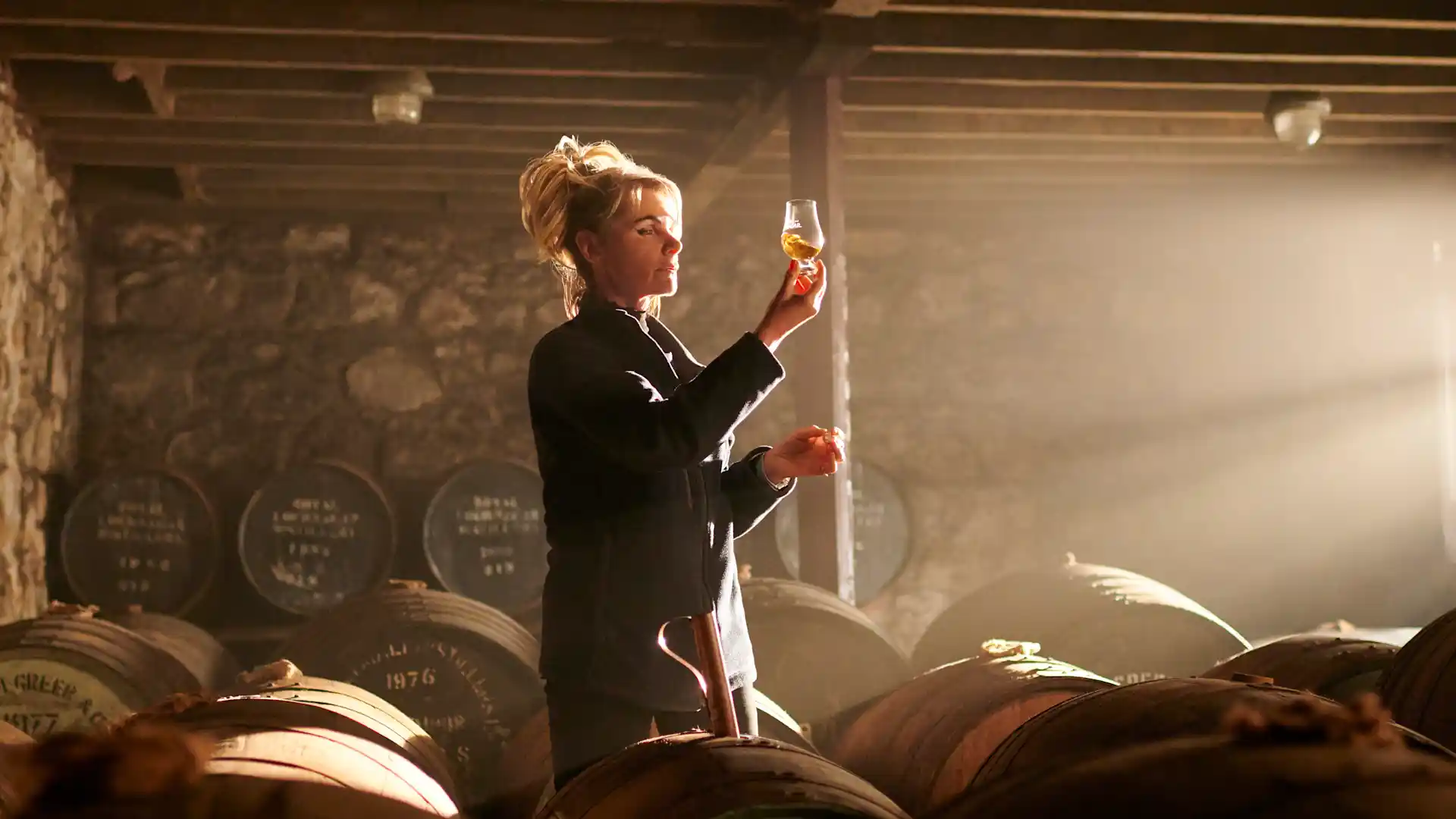 A tour guide from Royal Lochnagar holds a glass of whisky up to the light. She stands in a stone room filled with whisky barrels