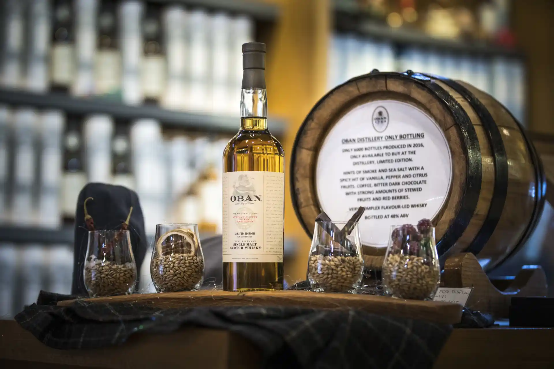 A bottle of Oban whisky sits on a wooden table surrounded by dram glasses containing barley and other assorted raw ingredients.