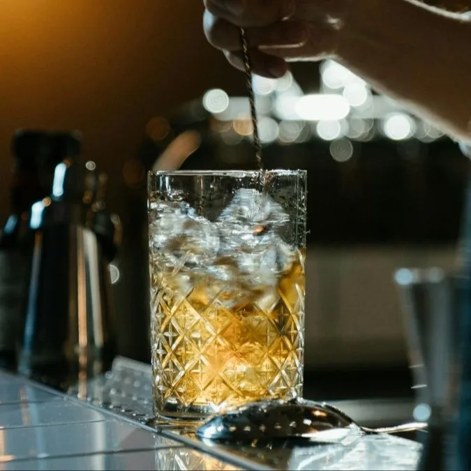 A person stirs whisky in a mixing glass filled with ice on a bar top