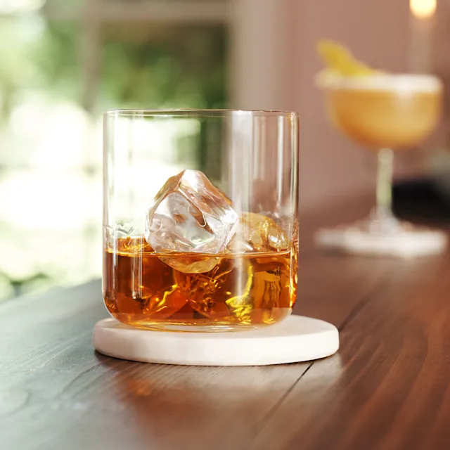 Op een bar staat een glas met whisky en ijs, met daarachter een cocktailshaker en een jigger.