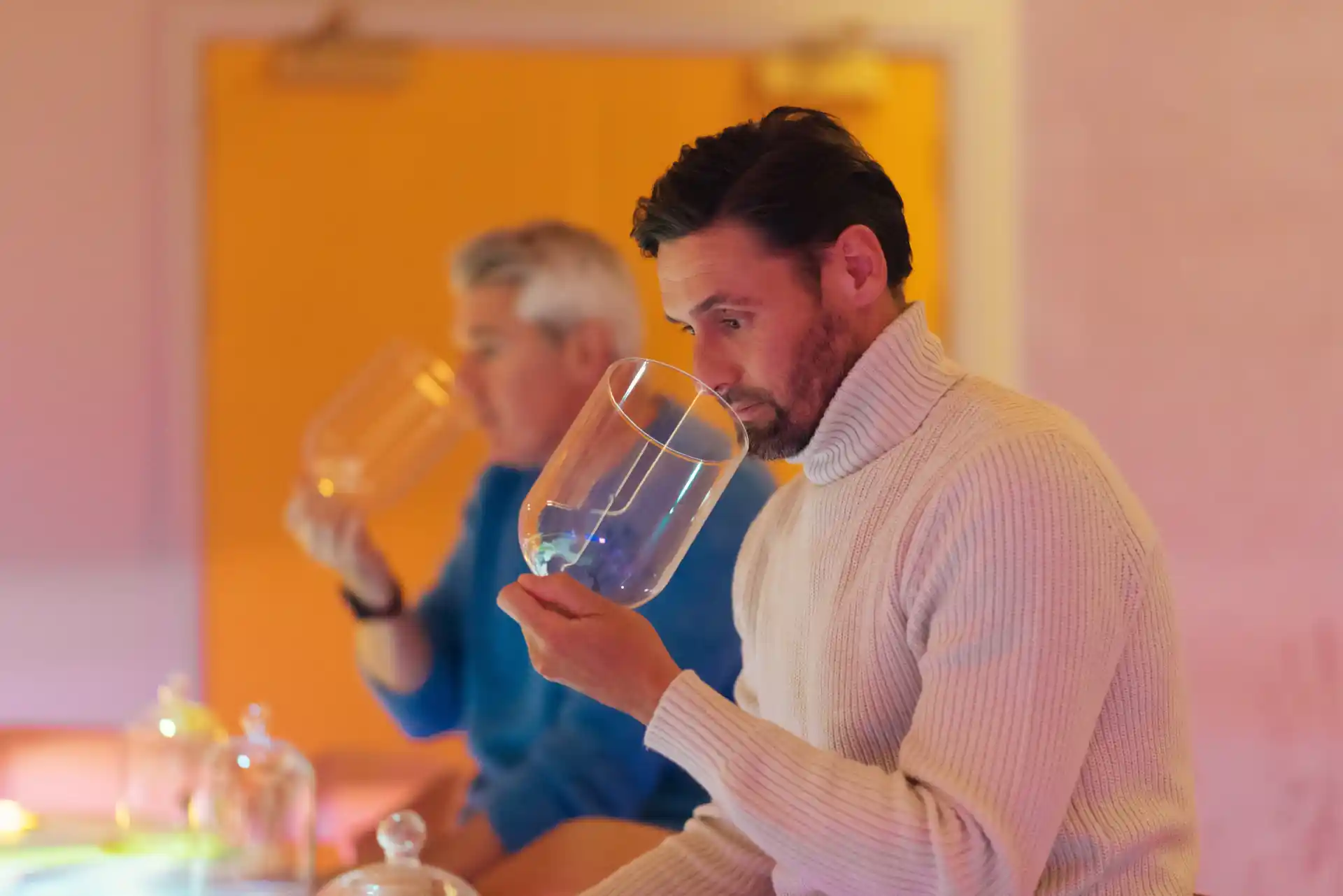  Two men sit at a table, holding glass cloches to their noses.