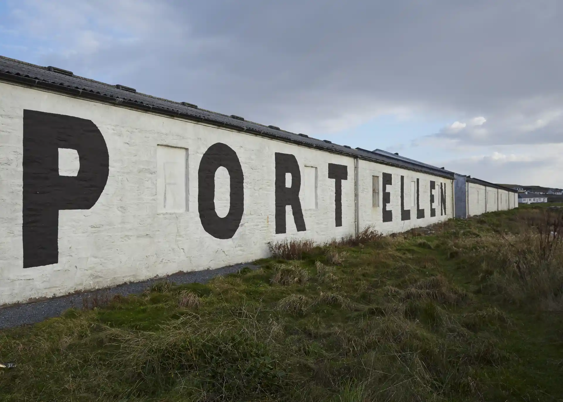 Voor de witte muren van het gebouw van de distilleerderij van Port Ellen ligt een grasveld. Op één muur staan in grote zwarte letters de woorden 'Port Ellen',