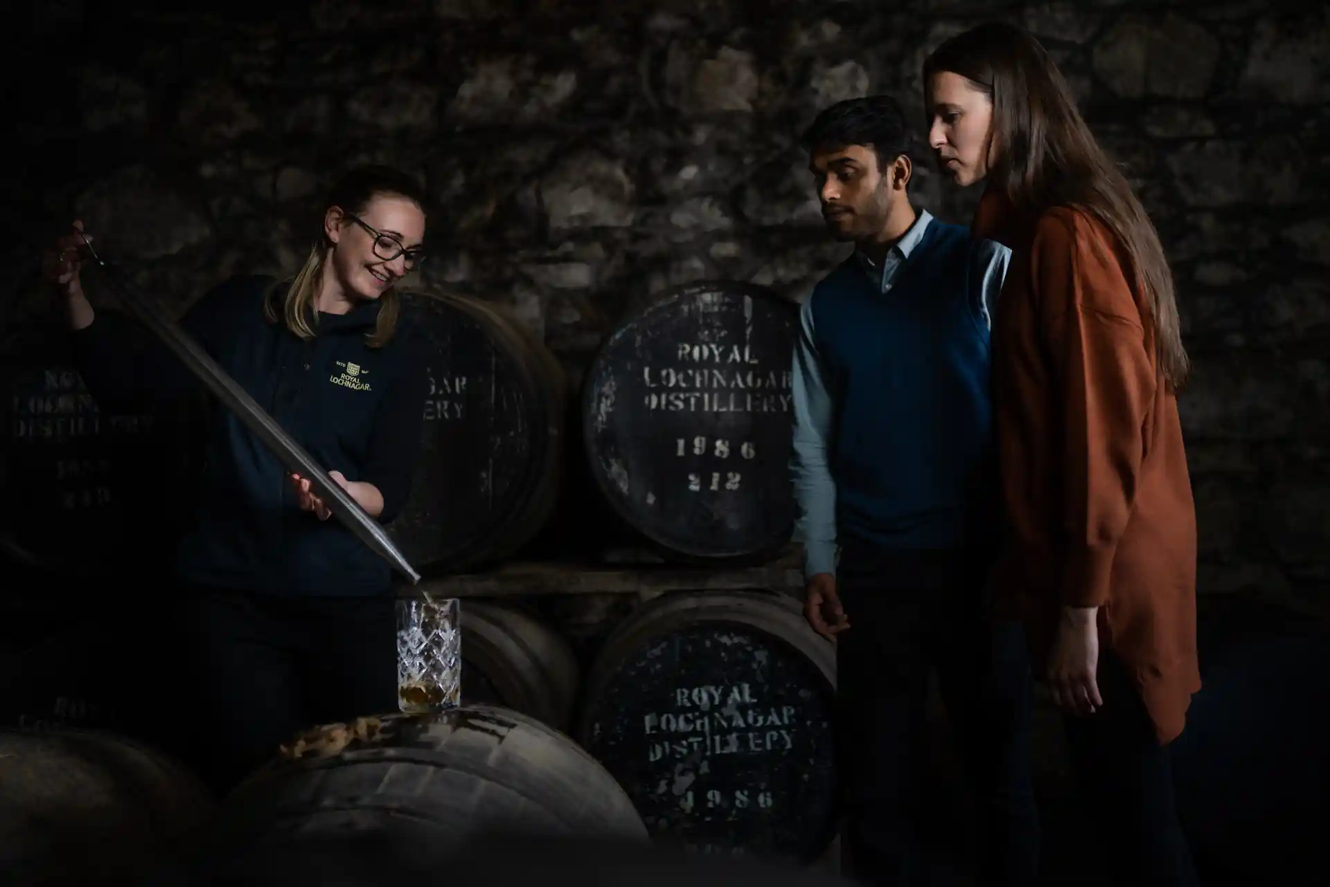 A Royal Lochnagar employee uses a large metal pipette to extract whisky from a wooden barrel and place it into a large glass decanter in front of two guests. Behind her are several wooden barrels laying on the sides, stacked on shelves.