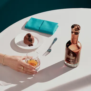 The Singleton bottle with fig and chocolate ganache dessert, a glass of whiskey, and a teal napkin on a white table.