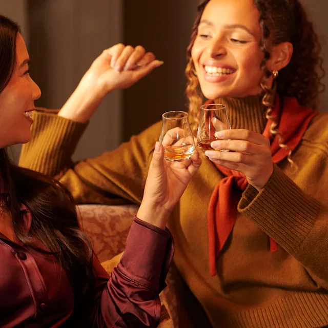 Two female friends sit on a sofa, enjoying whisky drams. They are smiling at each other.