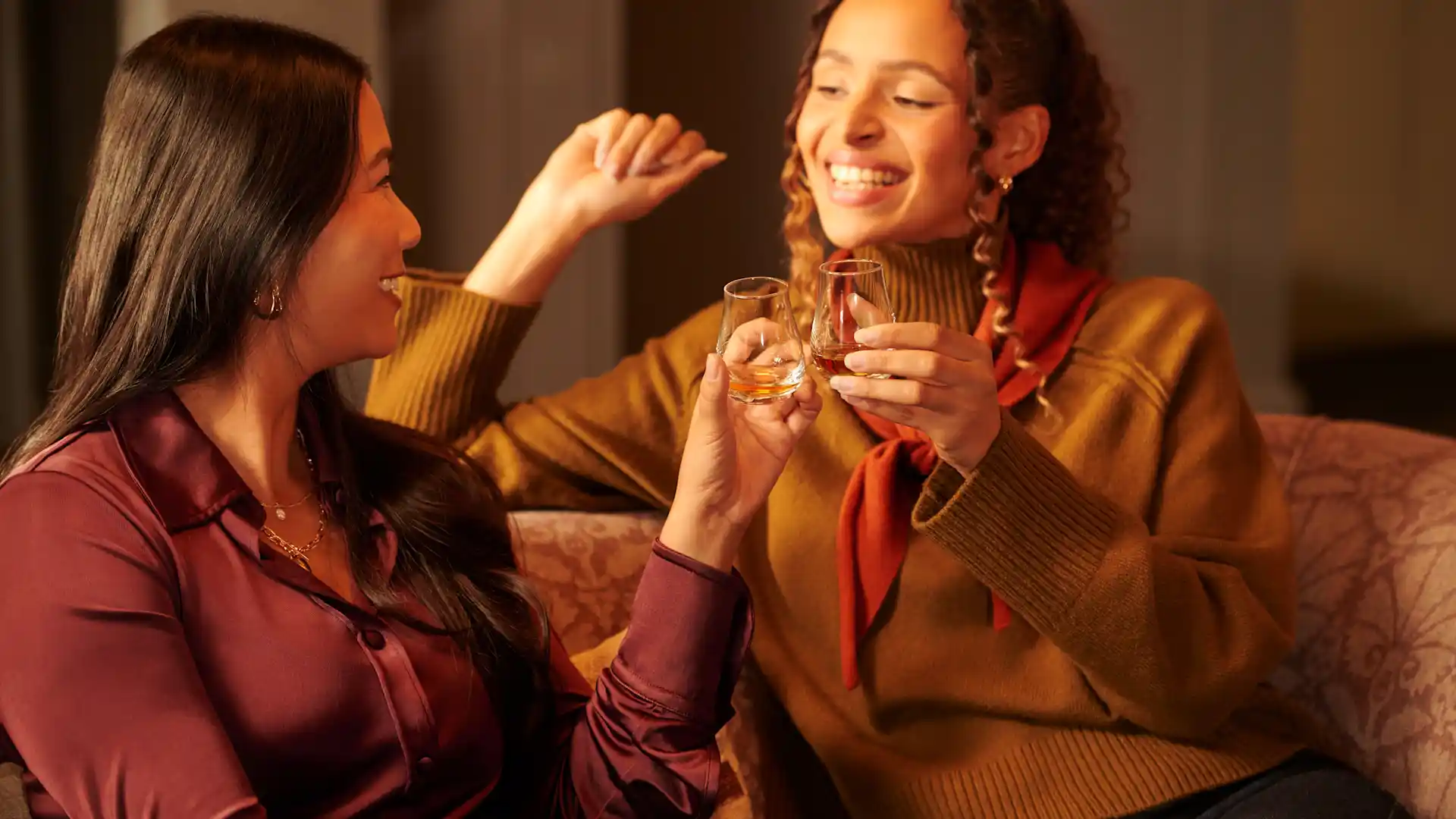 Two female friends sit on a sofa, enjoying whisky drams. They are smiling at each other.