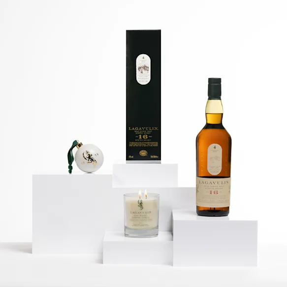 Lagavulin 16 Year Old whisky bottle with a matching box, a festive bauble, and a double-wick whisky-scented candle on white display blocks.
