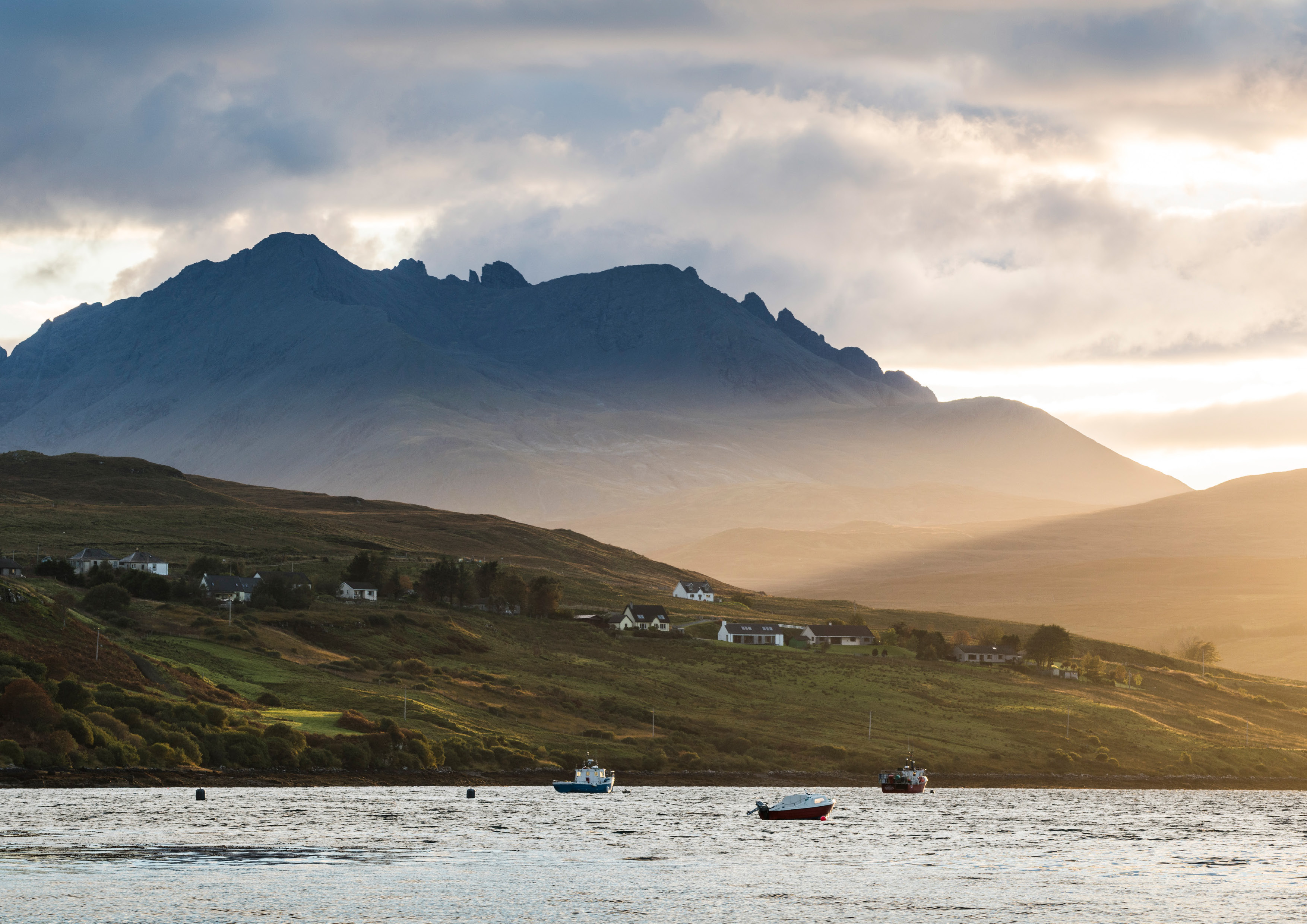 A Scottish loch, surrounded by hills and mountains which are covered in wispy clouds. Little boats bob on the loch.