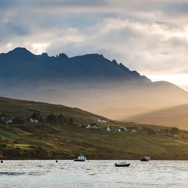 A Scottish loch, surrounded by hills and mountains which are covered in wispy clouds. Little boats bob on the loch.