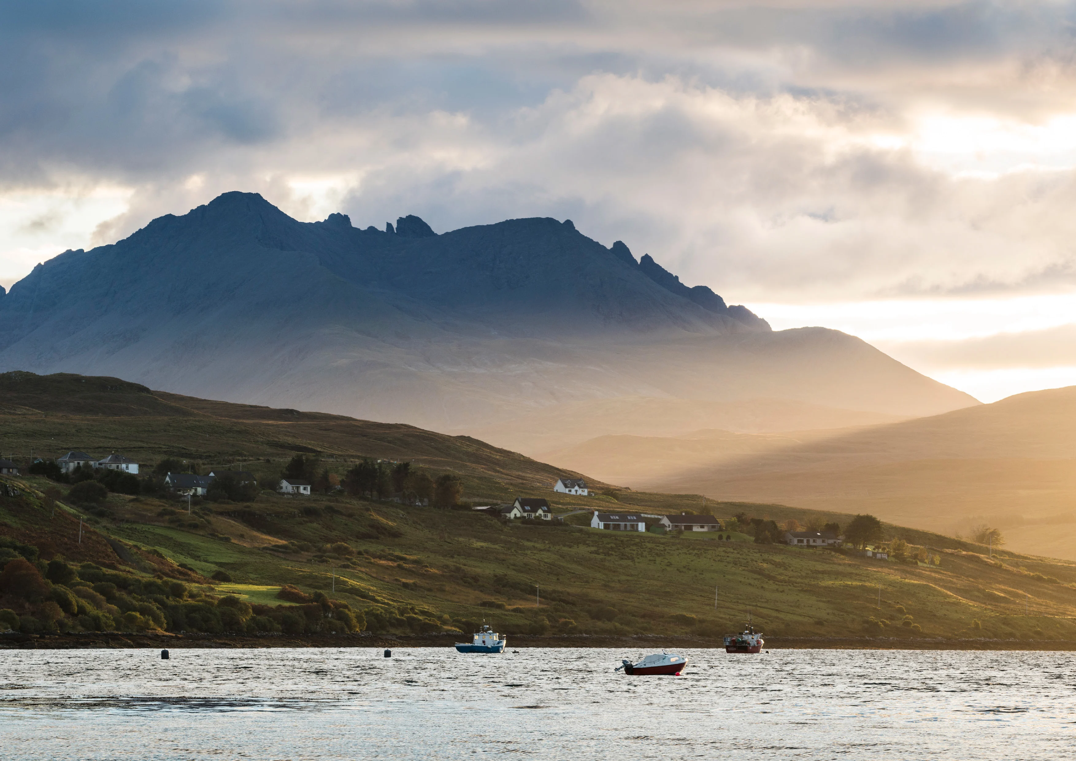 A Scottish loch, surrounded by hills and mountains which are covered in wispy clouds. Little boats bob on the loch.