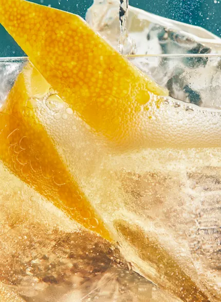 A close-up of a whisky cocktail containing ice and a lemon twist, with a bubbly liquid being poured into the glass.