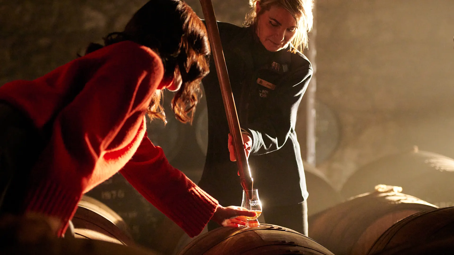 Een vrouw tapt whisky uit een houten vat met behulp van een metalen pipet. Ze giet de whisky in een proefglas dat wordt vastgehouden door een vrouw in een rode trui, die het glas bovenop het vat houdt.
