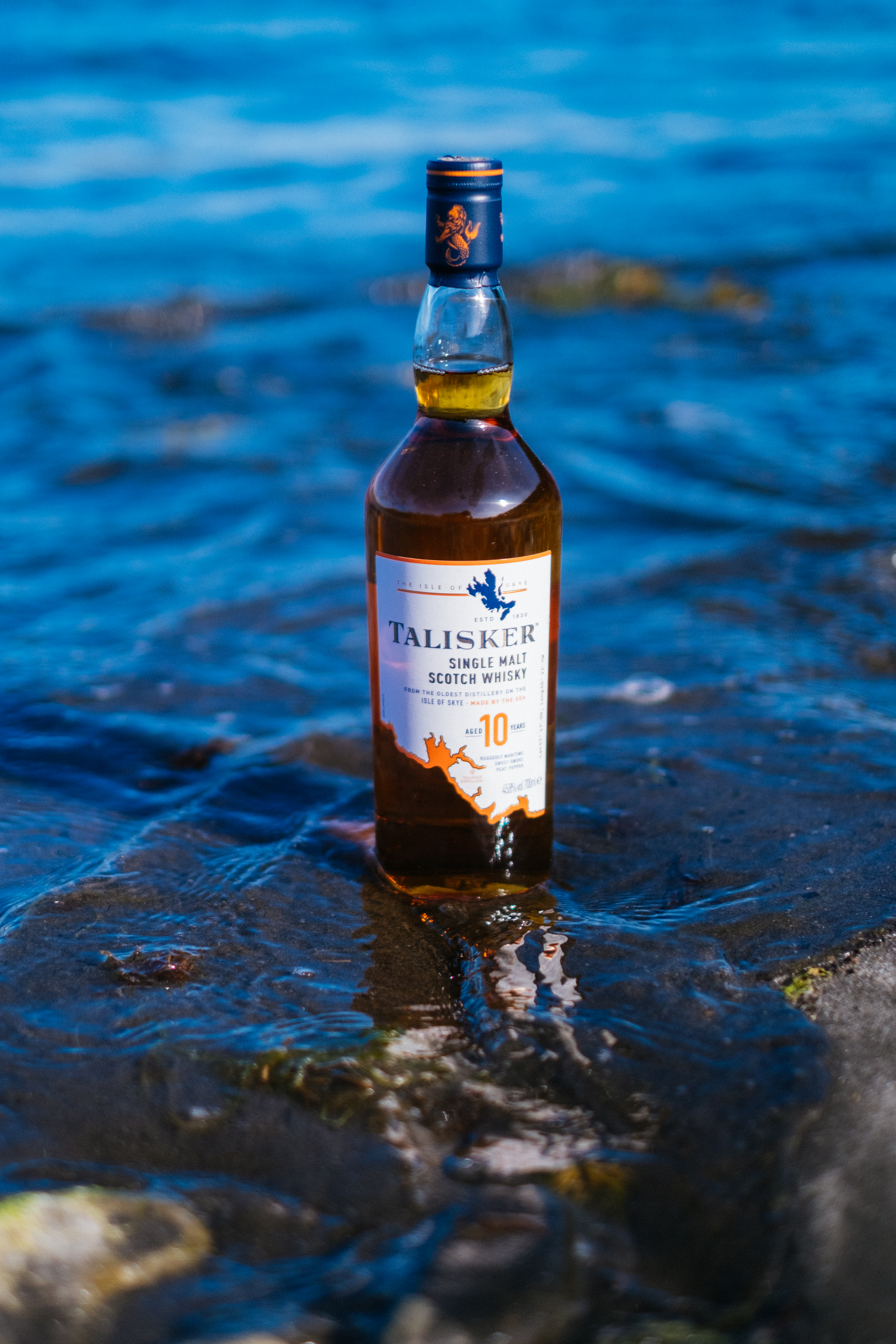 A bottle of Talisker 10 year old whisky sits on a a shoreline with the sea behind it