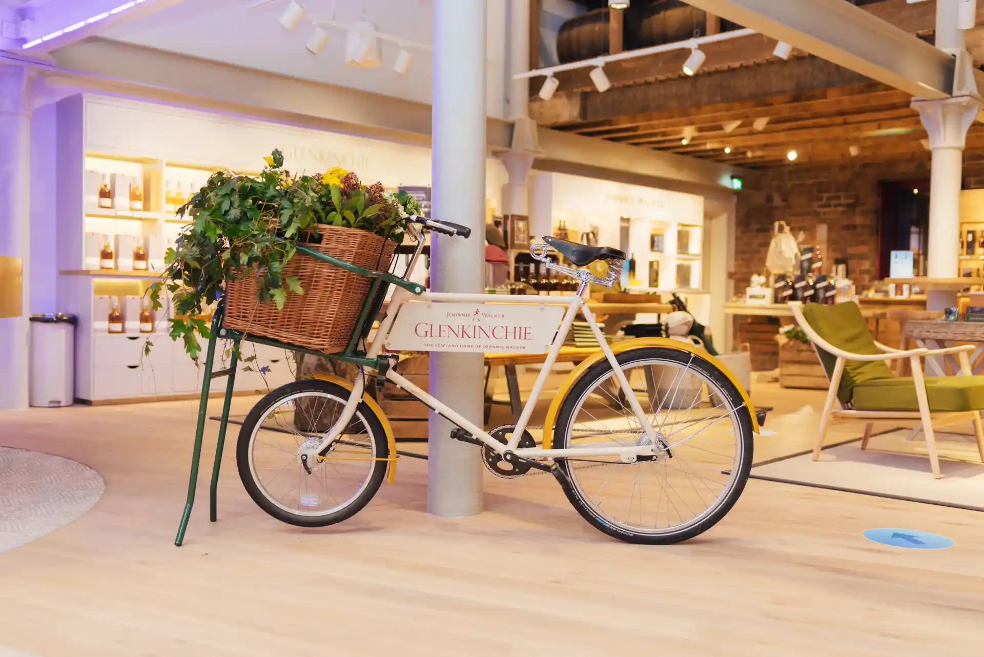  A white bicycle engraved with the words ‘Glenkinchie’ sits within a gift shop. It has a wicker basket from which several green plants spill out. Behind the bike are shelves containing assorted whisky bottles.