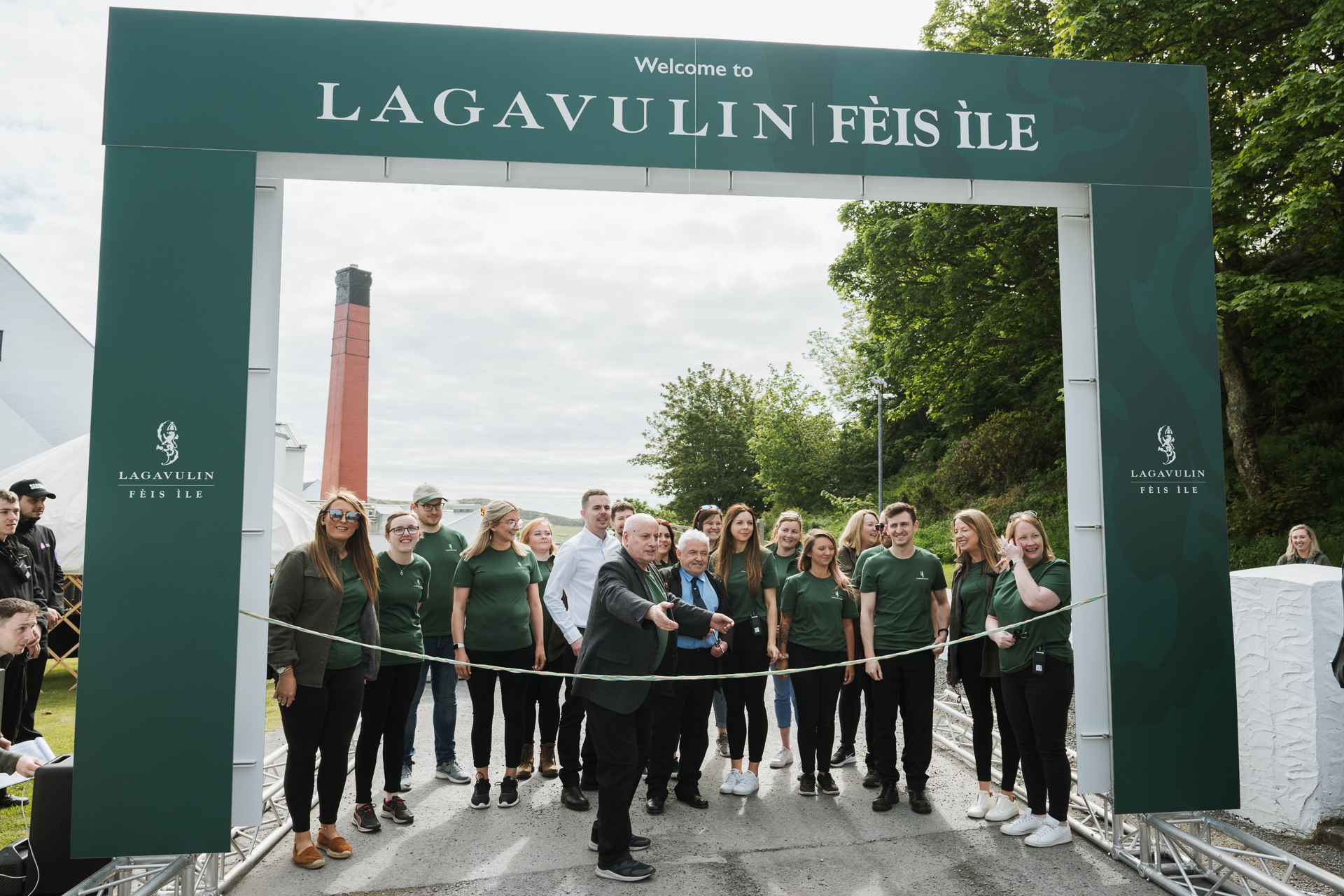 Eine Gruppe von Menschen, die hinter einem grünen Tor stehen, wobei einer die Linie durchschneidet und so symbolisch das Fèis ìle Festival eröffnet.