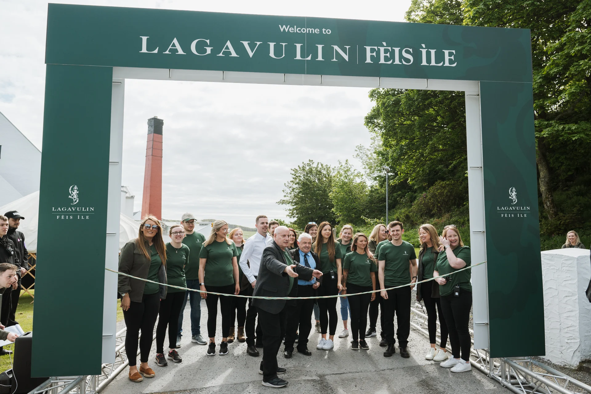 Eine Gruppe von Menschen, die hinter einem grünen Tor stehen, wobei einer die Linie durchschneidet und so symbolisch das Fèis ìle Festival eröffnet.
