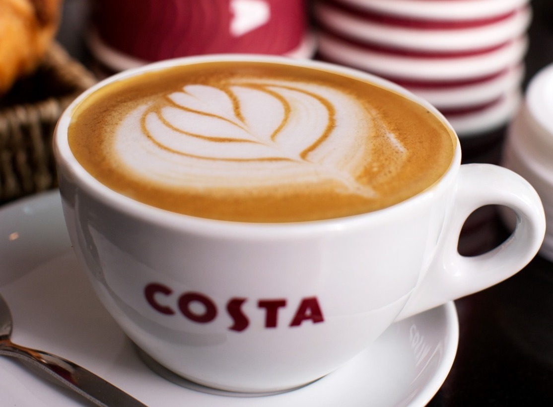 Costa Flat White in Porzellantasse mit Costa Pappbechern im Hintergrund