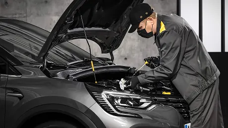 Une photographie montre un mécanicien effectuant l'entretien d'une voiture grise dont le capot est relevé. Le mécanicien, vêtu d'un masque noir, d'une casquette noire, de gants noirs et d'un uniforme gris à touches jaunes, est penché sur le compartiment moteur et vérifie le niveau d'huile à l'aide d'une jauge. Le bloc optique moderne et la calandre de la voiture sont visibles au premier plan.