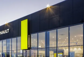 Une photographie d'architecture d'une concession automobile Renault moderne au crépuscule. Le bâtiment présente une façade supérieure sombre et texturée, éclairée par de petits spots, de larges baies vitrées laissant entrevoir un intérieur très lumineux, ainsi que des logos Renault en forme de losange bien visibles, accompagnés du nom « RENAULT » en blanc. Un haut pilier vertical d'un jaune vif — couleur emblématique de la marque — surplombe l'entrée principale, tandis qu'un totem indépendant de la même couleur se dresse à gauche du bâtiment.