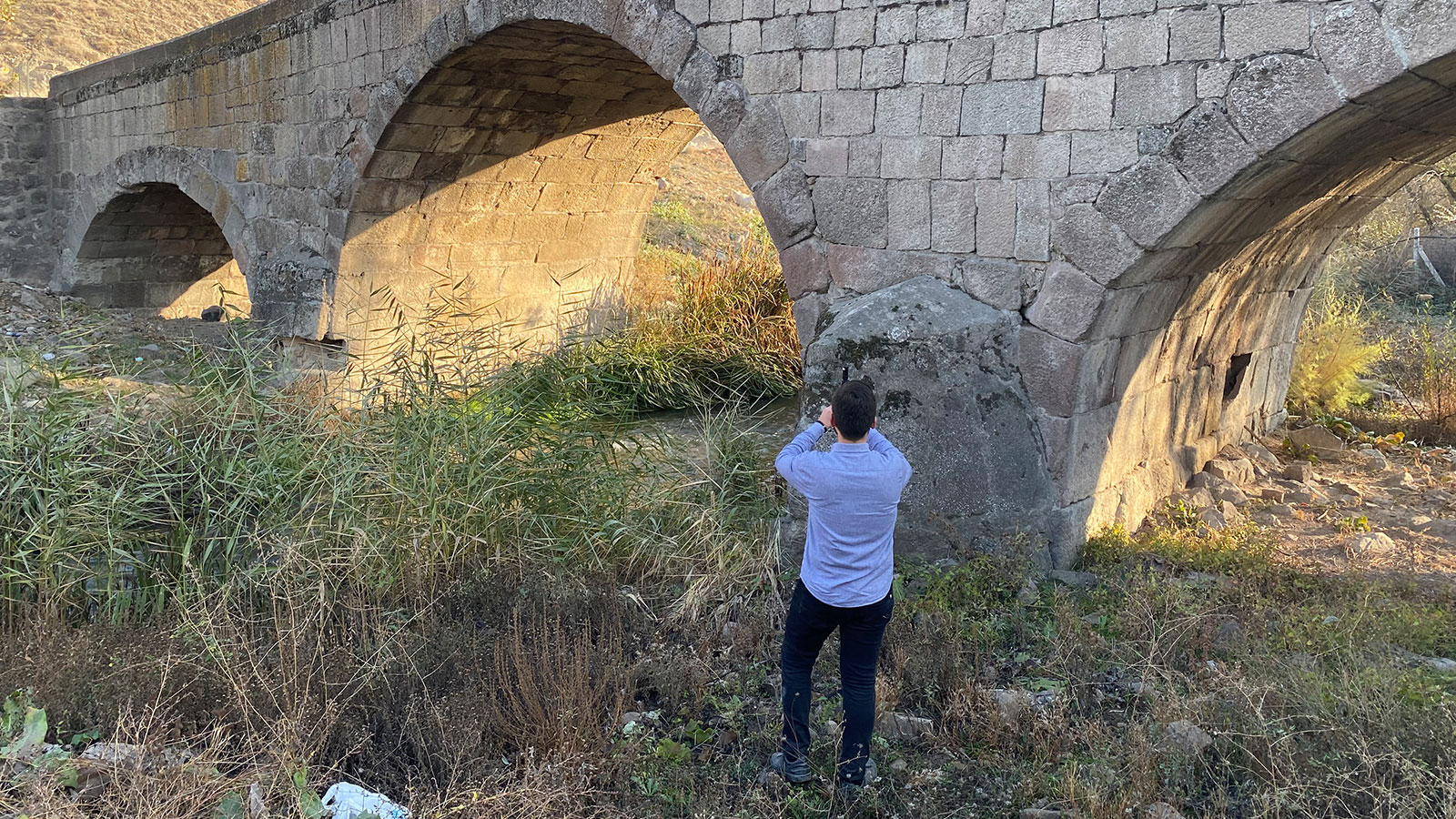 A person standing pointing the viDoc RTK rover at a stone bridge