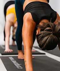 Woman in black workout clothes doing yoga on a mat, with another person blurred in the background.