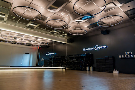 CorePower Yoga studio with mirrored wall, wood floor, circular ceiling lights, and blue neon “Flow SWEAT Savasana” sign