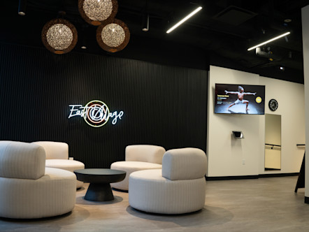 Modern CorePower Yoga East Village studio lounge with white chairs, neon "East Village" sign, woven pendant lights, and a screen showing the CPY FROI
