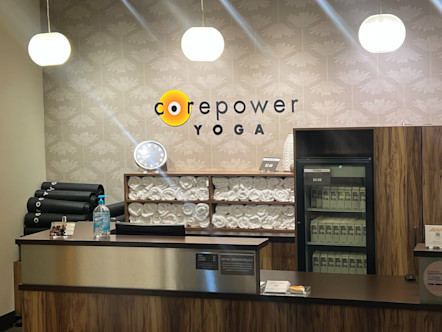 Large wooden and metal tiered front desk in front of a large CorePower Yoga sign centered on a grey and cream patterned wallpapered wall underneath three white orb light fixtures