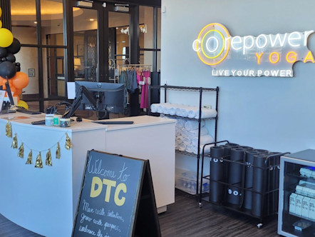 DTC front desk area with a neon CorePower Yoga sign on a grey wall behind a large white desk and towel racks below
