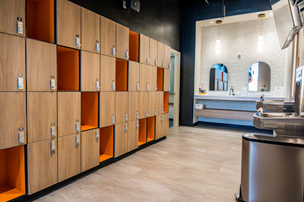 Modern locker room with wood lockers and orange cubbies, mirrors, sinks, and water fountains under pendant lights