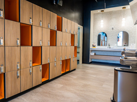 Modern locker room with wood lockers and orange cubbies, mirrors, sinks, and water fountains under pendant lights