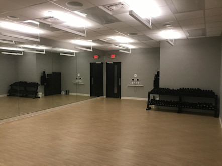 CPY studio room with grey walls and floor to ceiling mirrors on the front wall, light tan floors, and a black weight rack on the center of the back wall