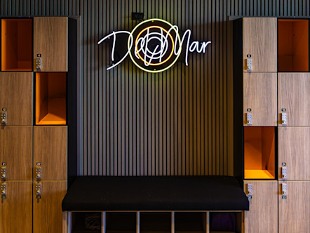 Neon “Del Mar” sign above a bench, flanked by numbered wooden lockers, with large woven pendant lights overhead.