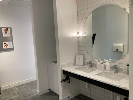 Grey tiled floors with white tiled walls and a large circular mirror above two sink spaces on a white marbled countertop vanity